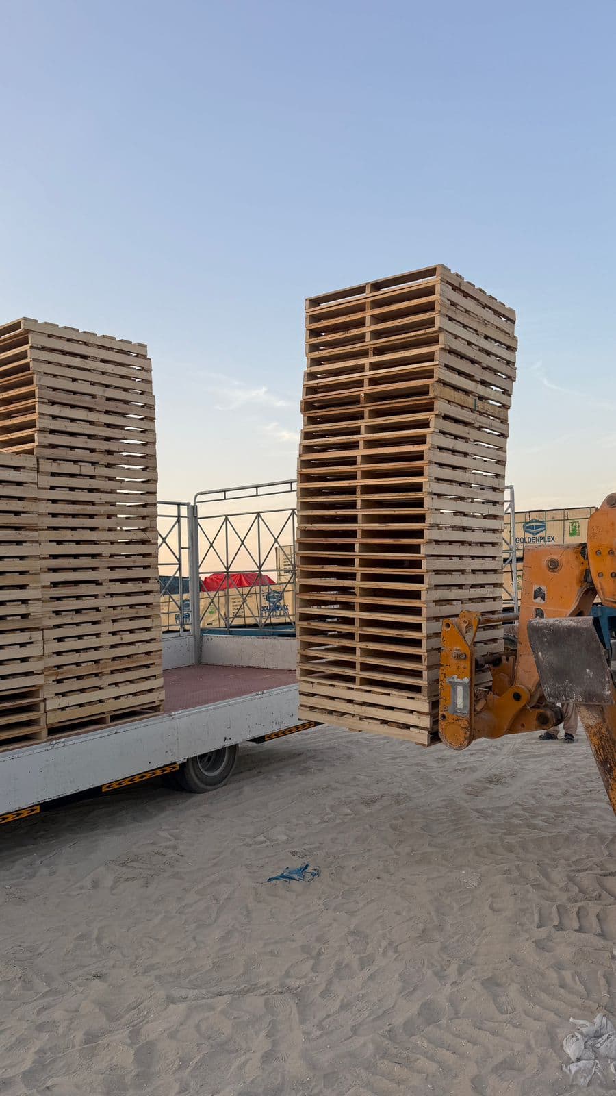 A telehandler lifting a stack of wooden pallets onto a flatbed truck at the Sharjah yard