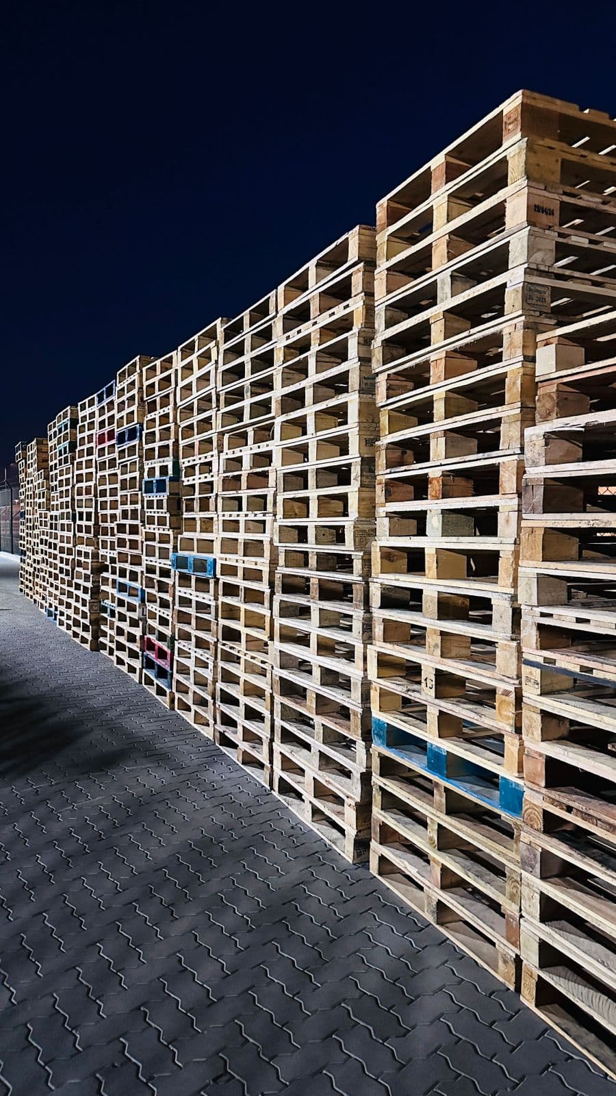 A long wall of used wooden pallets stacked in the yard, lit at night